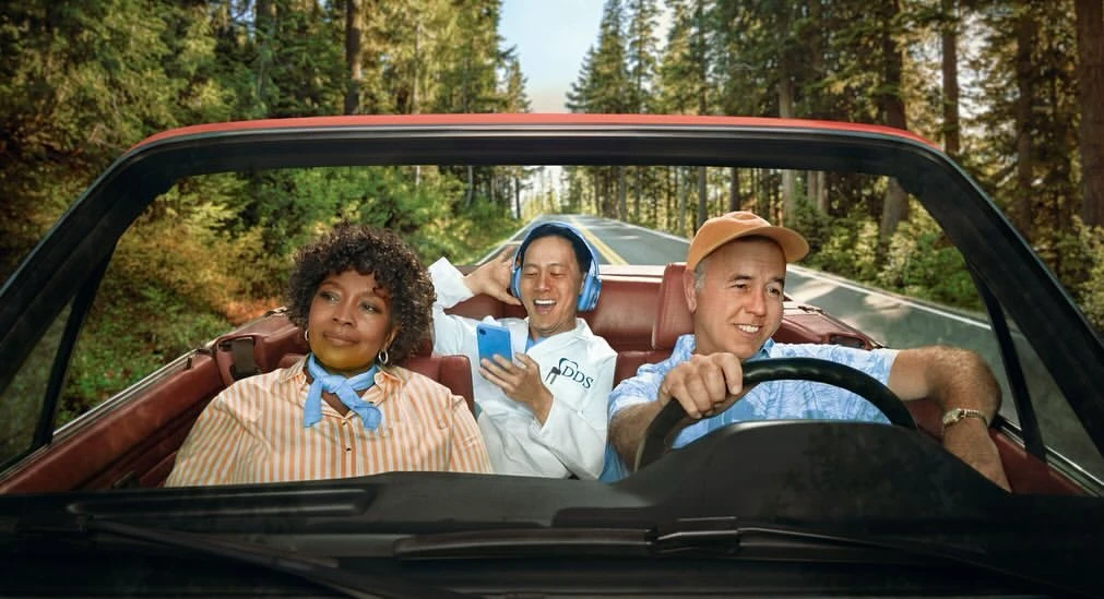 Three people ride in a red convertible car on a road lined with tall trees, creating a vibrant and scenic travel image.